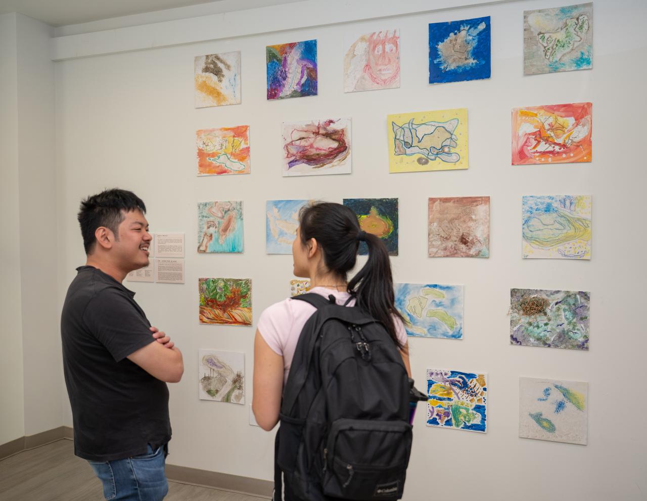 Two people stand facing each other beside a gallery wall displaying colorful abstract artworks arranged in rows.
