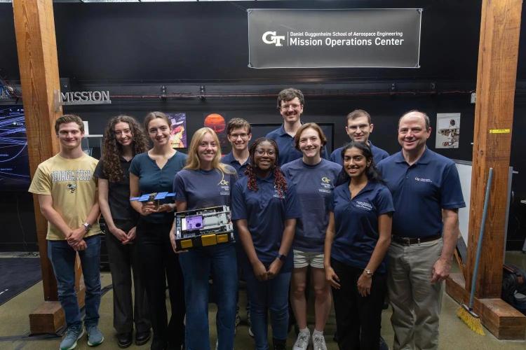 Georgia Tech students, faculty, and staff pose with spacecraft hardware beneath the Daniel Guggenheim School of Aerospace Engineering Mission Operations Center sign.